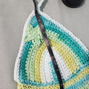 Pu&ograve; includere: Un top all'uncinetto triangolare con strisce orizzontali nei toni del verde acqua, verde lime e bianco. Un metro a nastro nero &egrave; posizionato verticalmente sopra il centro della coppa, indicando un'altezza di circa 16,5 cm.