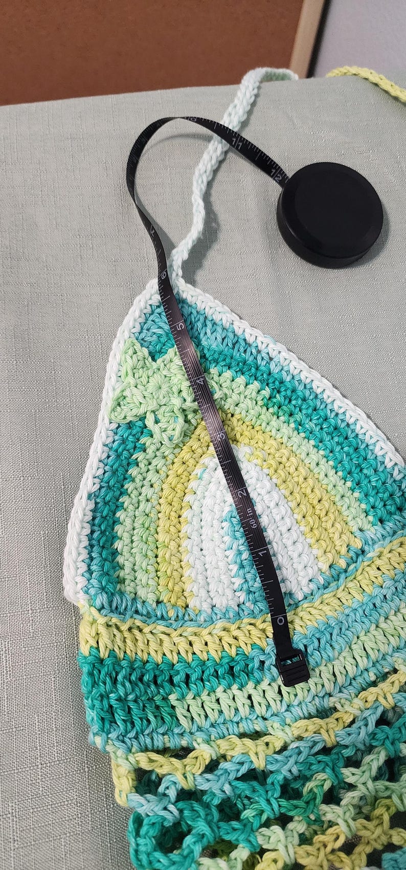 Pu&ograve; includere: Un top all'uncinetto triangolare con strisce orizzontali nei toni del verde acqua, verde lime e bianco. Un metro a nastro nero &egrave; posizionato verticalmente sopra il centro della coppa, indicando un'altezza di circa 16,5 cm.