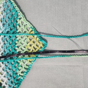 Pu&ograve; includere: Un top triangolare all'uncinetto con un motivo variegato di filato color verde acqua, verde lime e bianco. Il capo include lunghi lacci che terminano con una forma a stella e a foglia, con un metro a nastro nero che indica 38 centimetri.