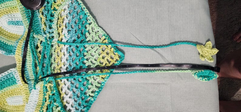 Pu&ograve; includere: Un top triangolare all'uncinetto con un motivo variegato di filato color verde acqua, verde lime e bianco. Il capo include lunghi lacci che terminano con una forma a stella e a foglia, con un metro a nastro nero che indica 38 centimetri.