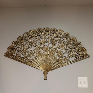 May include: A large, gold-colored wall-mounted fan decoration featuring an intricate, openwork filigree design with floral and scroll motifs. The fan is shaped in a wide, semi-circular arc with a detailed handle at the base, displaying a metallic finish.