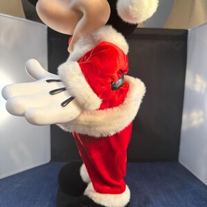 May include: A side view of a Mickey Mouse figurine dressed in a Santa Claus outfit. The red velvet suit is trimmed with white faux fur. The figurine has white gloves, black shoes, and a Santa hat with a white pom-pom.