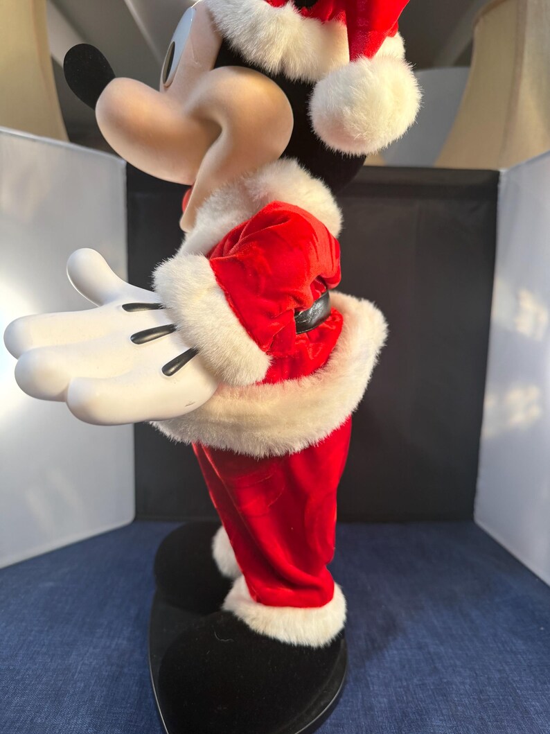 May include: A side view of a Mickey Mouse figurine dressed in a Santa Claus outfit. The red velvet suit is trimmed with white faux fur. The figurine has white gloves, black shoes, and a Santa hat with a white pom-pom.