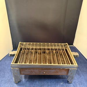 Puede incluir: Una antigua parrilla eléctrica rectangular con una rejilla metálica y elementos calefactores. La parrilla tiene un marco azul-gris con un cajón marrón y asas metálicas. Las patas también son azul-gris.
