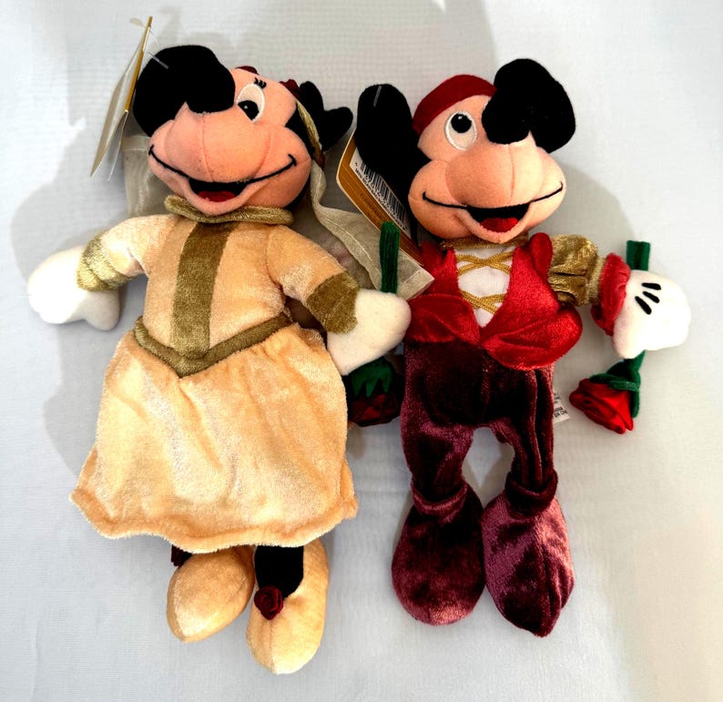 May include: Two plush Disney character dolls. One doll is dressed in a peach-colored dress with gold accents, and the other doll is wearing a red and burgundy outfit. Both dolls have black ears, pink faces, and white gloves. Each doll is holding a small green and red item.