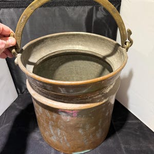May include: A copper bucket with a brass handle. The bucket has a cylindrical shape with a slightly flared rim. The handle is arched and attached to the bucket with decorative brackets. The copper has a patina, showing signs of age and use.