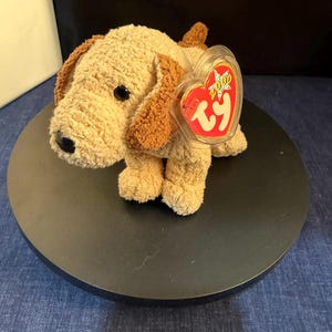 Könnte beinhalten: Ein Stoffhund-Spielzeug mit hellbraunem und braunem Fell, schwarzen Augen und einem roten herzförmigen Anhänger mit der Aufschrift "2000 TY". Das Stofftier befindet sich auf einer schwarzen, kreisförmigen Oberfläche.