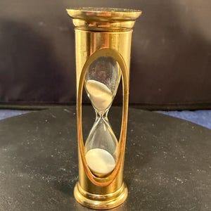 May include: A brass hourglass with a clear glass center containing white sand. The hourglass has a decorative, oval-shaped frame and a cylindrical body. The top and bottom are also brass, with a decorative edge. The hourglass is a classic timekeeping device.