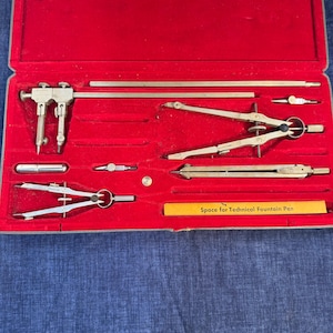 以下が含まれることがあります： 赤色のベルベットの裏地が付いたケースに入った、ヴィンテージの製図セット。セットには、シルバー色のコンパス、ディバイダー、その他の製図ツールが含まれています。「Space for Technical Fountain Pen」と書かれた黄色のラベル。