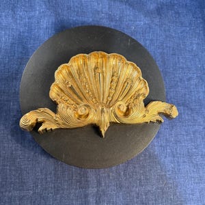 Puede incluir: Un objeto decorativo de metal dorado con forma de concha, con intrincados detalles en relieve y volutas en la base, montado sobre un disco negro circular. La concha presenta un patrón en forma de abanico con crestas verticales y adornos.