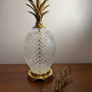 Puede incluir: Una lámpara de mesa vintage con forma de piña. La lámpara presenta un cuerpo de vidrio transparente tallado en diamante y una base y parte superior de metal dorado. El diseño de la lámpara incluye una corona de piña detallada y un cable con enchufe.