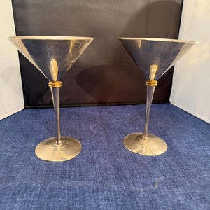 Puede incluir: Dos copas de martini plateadas con una banda dorada. Las copas tienen una forma de cuenco triangular clásica y un tallo largo y delgado. La base de cada copa es redonda. Altura aproximada de 15 cm.