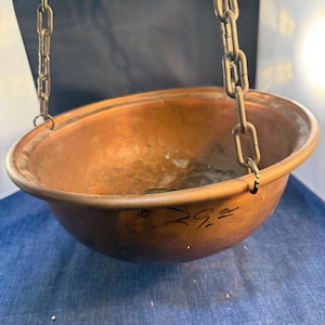 May include: A round copper bowl with a smooth, metallic finish and a wide rim. The bowl is suspended by three metal chains attached to the outer edge. The exterior of the bowl features handwritten black text that appears to read 29.00.