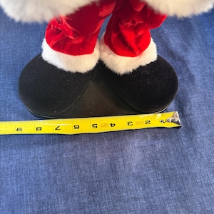 May include: Close-up of a Santa Claus figurine's legs and feet. The red velvet pants are trimmed with white faux fur. The black shoes are approximately 8 inches long, measured with a yellow tape measure. The figurine is on a blue surface.