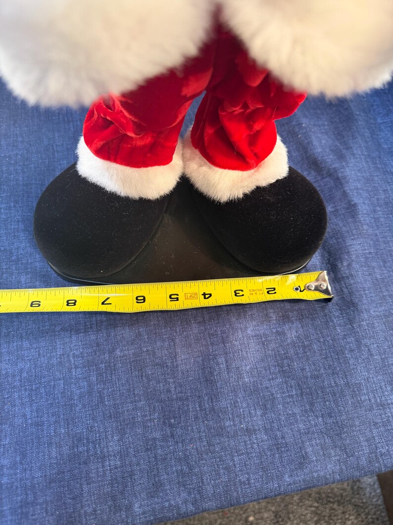 May include: Close-up of a Santa Claus figurine's legs and feet. The red velvet pants are trimmed with white faux fur. The black shoes are approximately 8 inches long, measured with a yellow tape measure. The figurine is on a blue surface.
