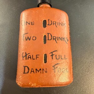 May include: A brown leather flask with a black cap. The flask has black text that reads "ONE DRINK", "TWO DRINKS", "HALF FULL", and "DAMN FULL". The flask has a rectangular shape.