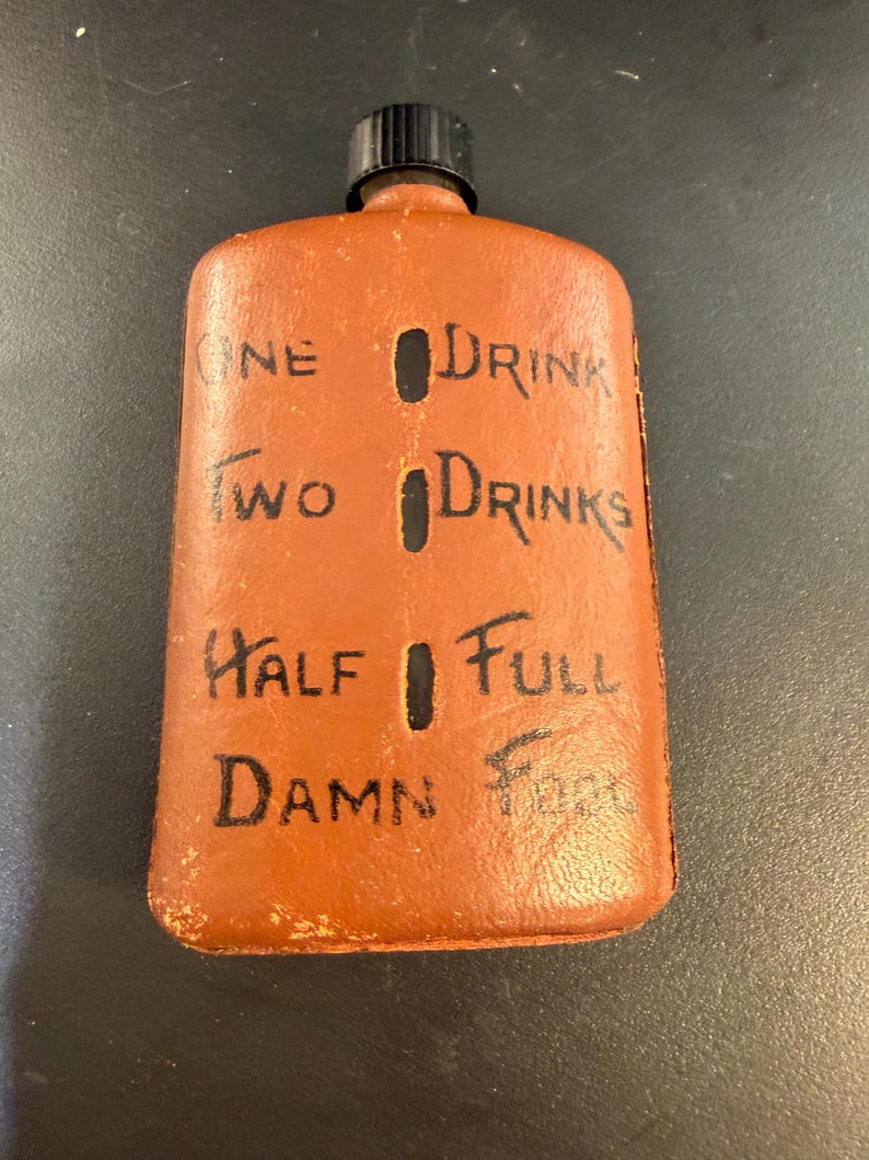 May include: A brown leather flask with a black cap. The flask has black text that reads "ONE DRINK", "TWO DRINKS", "HALF FULL", and "DAMN FULL". The flask has a rectangular shape.