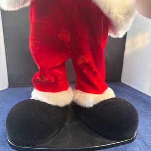 May include: A festive Santa Claus figure, featuring red velvet pants with white faux fur trim and large black shoes. The figure's upper body is adorned with white faux fur. The figure is standing on a black base, perfect for holiday decor.