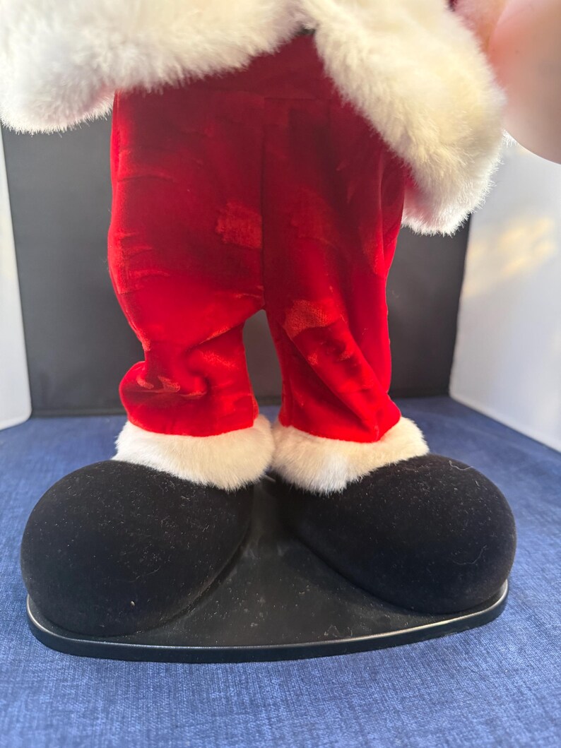 May include: A festive Santa Claus figure, featuring red velvet pants with white faux fur trim and large black shoes. The figure's upper body is adorned with white faux fur. The figure is standing on a black base, perfect for holiday decor.
