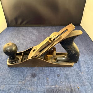 Puede incluir: Un cepillo de mano vintage, con un cuerpo de madera oscura y un mango y pomo negros. La cuchilla metálica y el mecanismo de ajuste muestran signos de desgaste. Esta herramienta de carpintería está diseñada para dar forma y alisar superficies de madera.