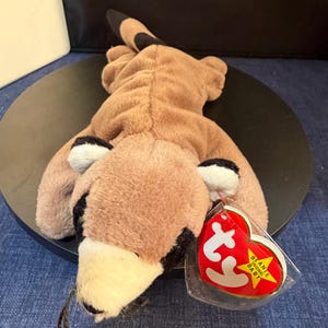 Puede incluir: Un juguete de peluche de mapache marrón y beige con detalles en negro en las orejas y la cola. El juguete tiene un hocico color crema y una etiqueta roja en forma de corazón que dice "Beanie Baby".