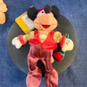May include: A plush Mickey Mouse doll wearing a red vest, burgundy pants, and a red and gold hat. The doll has black ears, white gloves, and is holding a small red strawberry. A yellow tag is attached.