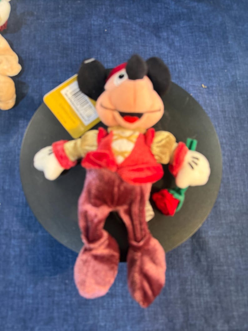 May include: A plush Mickey Mouse doll wearing a red vest, burgundy pants, and a red and gold hat. The doll has black ears, white gloves, and is holding a small red strawberry. A yellow tag is attached.