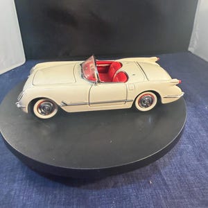 Peut inclure: Modèle réduit de voiture décapotable vintage crème avec intérieur rouge. La voiture a des pneus à flancs blancs et des accents chromés. Le modèle est présenté sur une plateforme circulaire noire, mettant en valeur son design et ses détails classiques.