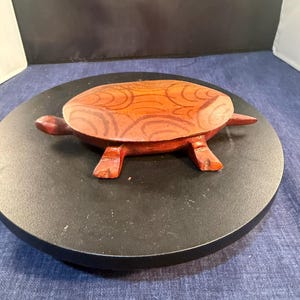 May include: A carved wooden turtle figurine with a reddish-brown finish. The shell features a darker brown, hand-painted pattern of concentric, curved lines. The turtle has a pointed tail, four short legs, and a small head extending from the body.