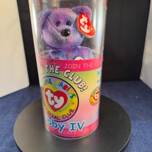 Può includere: Un peluche a forma di orsetto Ty Beanie Baby viola e blu all'interno di un contenitore di plastica trasparente. Il contenitore ha un'etichetta colorata con il testo "Join the Club!" e "Official Club". Un'etichetta rossa a forma di cuore è attaccata all'orsetto.