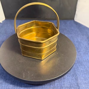 May include: A small, hexagonal brass basket featuring a polished metallic finish and a single arched handle attached to the sides. The container has a multi-tiered, ribbed exterior design with flat panels and a hollow, open top for storage or display.