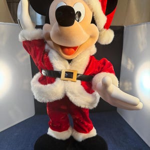 May include: A Mickey Mouse figure dressed in a Santa Claus outfit. The figure is wearing a red velvet suit with white faux fur trim, a black belt with a gold buckle, and a Santa hat. Mickey's black ears and shoes are visible.