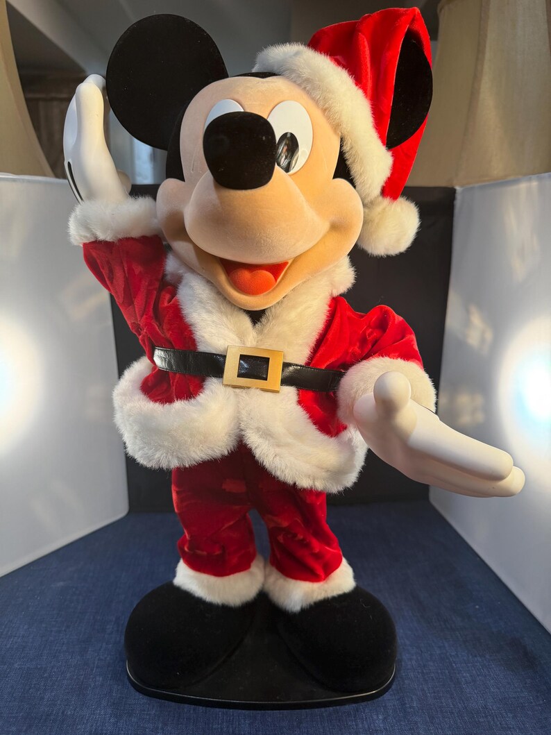 May include: A Mickey Mouse figure dressed in a Santa Claus outfit. The figure is wearing a red velvet suit with white faux fur trim, a black belt with a gold buckle, and a Santa hat. Mickey's black ears and shoes are visible.