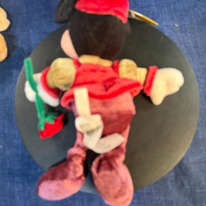 May include: A plush doll of a character wearing a red outfit, a matching hat, and white gloves. The doll has black hair and a painted face. It is holding a green and red item. The doll is on a dark surface.