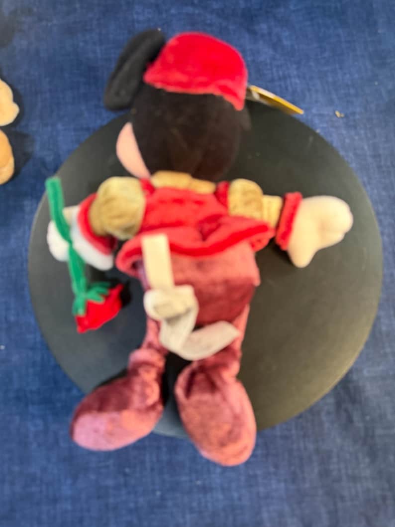May include: A plush doll of a character wearing a red outfit, a matching hat, and white gloves. The doll has black hair and a painted face. It is holding a green and red item. The doll is on a dark surface.