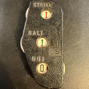 Op de afbeelding: Een zwarte Rawlings honkbal-scheidsrechterindicator met een getextureerd oppervlak. Het apparaat heeft drie ronde vensters die de cijfers 1, 1 en 0 weergeven in rood, groen en zwart, gelabeld met de witte tekst STRIKE, BALL en OUT, en zijwieltjes.