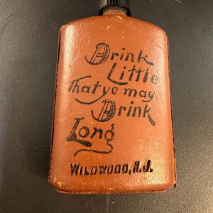 May include: A brown leather flask with a black cap. The flask has the words "Drink Little That ye may Drink Long" and "Wildwood, N.J." printed on it in black script.