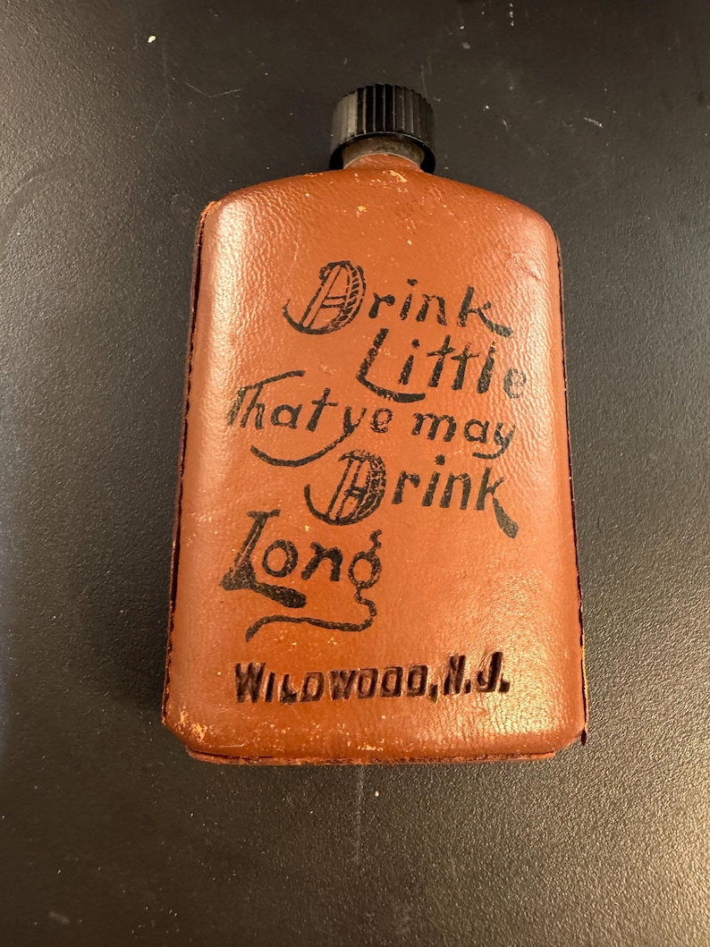 May include: A brown leather flask with a black cap. The flask has the words "Drink Little That ye may Drink Long" and "Wildwood, N.J." printed on it in black script.