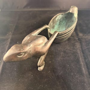 Könnte beinhalten: Eine bronzefarbene Skulptur einer vogelartigen Kreatur mit einem schalenförmigen Körper und einem echsenartigen Kopf und Armen. Das Innere der Schale hat eine grüne Patina. Die Skulptur befindet sich auf einer dunklen Oberfläche.