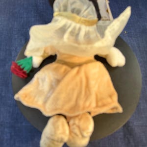 May include: A plush Minnie Mouse figurine, viewed from the back, wearing a light yellow dress and hat with a gold trim. The figure is holding a red and green item. The figurine is on a dark gray circular base.