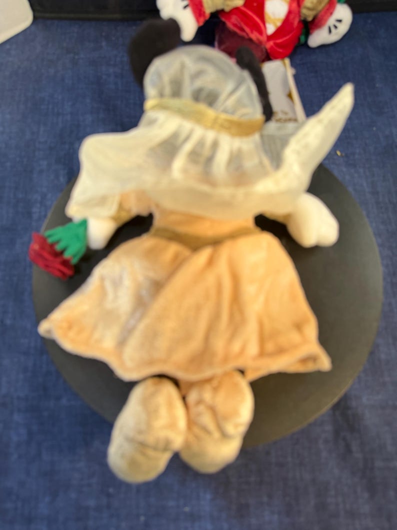 May include: A plush Minnie Mouse figurine, viewed from the back, wearing a light yellow dress and hat with a gold trim. The figure is holding a red and green item. The figurine is on a dark gray circular base.