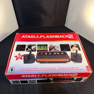 Op de afbeelding: Een Atari Flashback 2 klassieke spelcomputerdoos met een zwarte console met houtnerf en twee zwarte joysticks. De verpakking toont de tekst Atari Flashback 2, 40 ingebouwde spellen en titels zoals Pong, Asteroids en Pitfall.