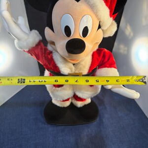 May include: A Mickey Mouse plush figure dressed as Santa Claus, wearing a red suit with white trim and a matching hat. The figure is approximately 12 inches tall, with outstretched arms.