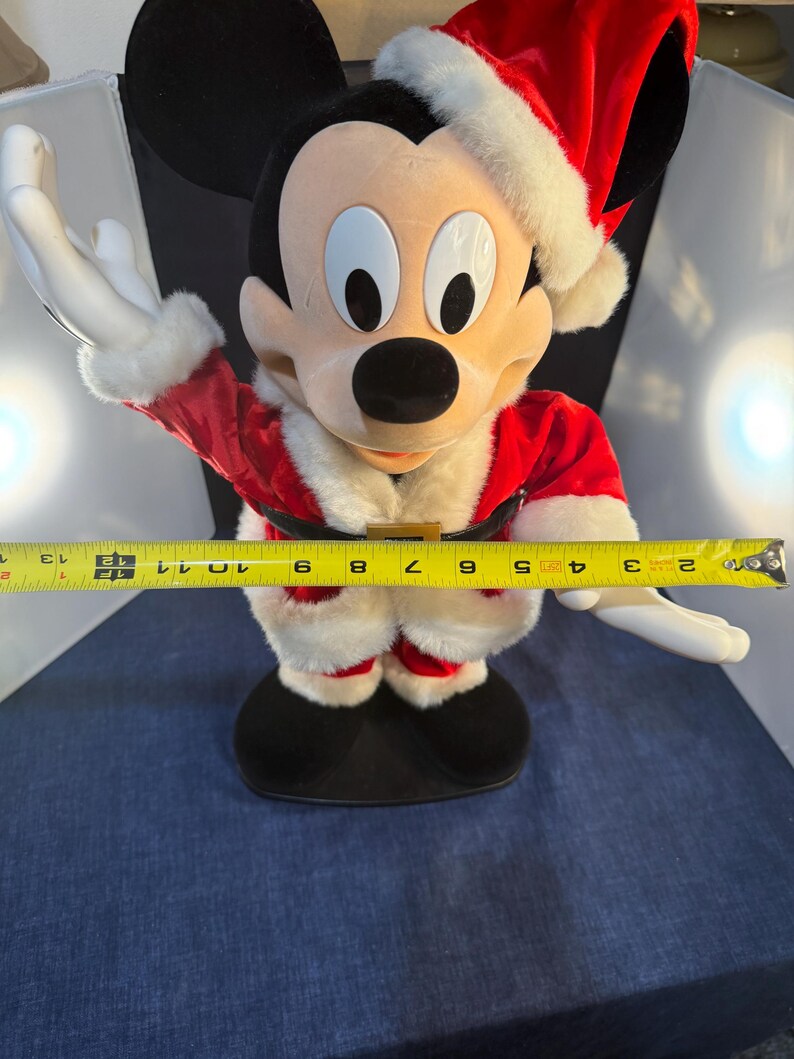 May include: A Mickey Mouse plush figure dressed as Santa Claus, wearing a red suit with white trim and a matching hat. The figure is approximately 12 inches tall, with outstretched arms.