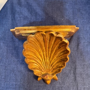May include: A gold-colored wall shelf featuring a prominent, carved scallop shell design as the support bracket. The shelf has a flat, rectangular top surface and a textured, metallic finish with visible highlights and shadows across the shell.