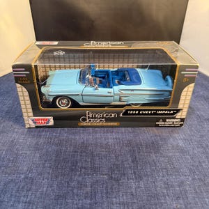 Op de afbeelding: Een 1:24 schaal die-cast model van een lichtblauwe 1958 Chevy Impala cabriolet met een donkerblauw interieur, verpakt in een rechthoekige kartonnen doos met het label American Classics en Motor Max met een doorzichtig plastic venster.
