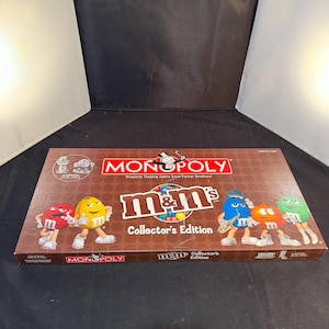 Op de afbeelding: Een rechthoekige Monopoly M&M's Collector's Edition bordspeldoos. De bruine doos bevat het Monopoly-logo, het M&M's-logo en illustraties van rode, gele, blauwe, oranje en groene M&M-personages. De tekst bevat Collector's Edition.
