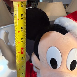 May include: A Mickey Mouse plush toy wearing a Santa hat, measured at approximately 16 inches tall. The plush has black ears, a red and white Santa hat, and a smiling face. The toy is a festive holiday decoration.