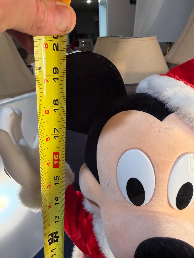 May include: A Mickey Mouse plush toy wearing a Santa hat, measured at approximately 16 inches tall. The plush has black ears, a red and white Santa hat, and a smiling face. The toy is a festive holiday decoration.