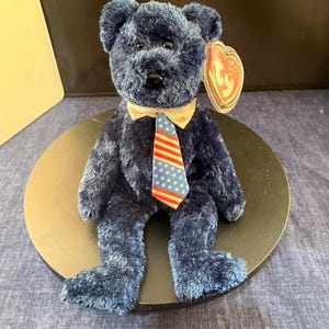 May include: A navy blue plush teddy bear with a cream-colored bow tie and a patriotic tie featuring stars and stripes. The bear has a tag attached to its ear.